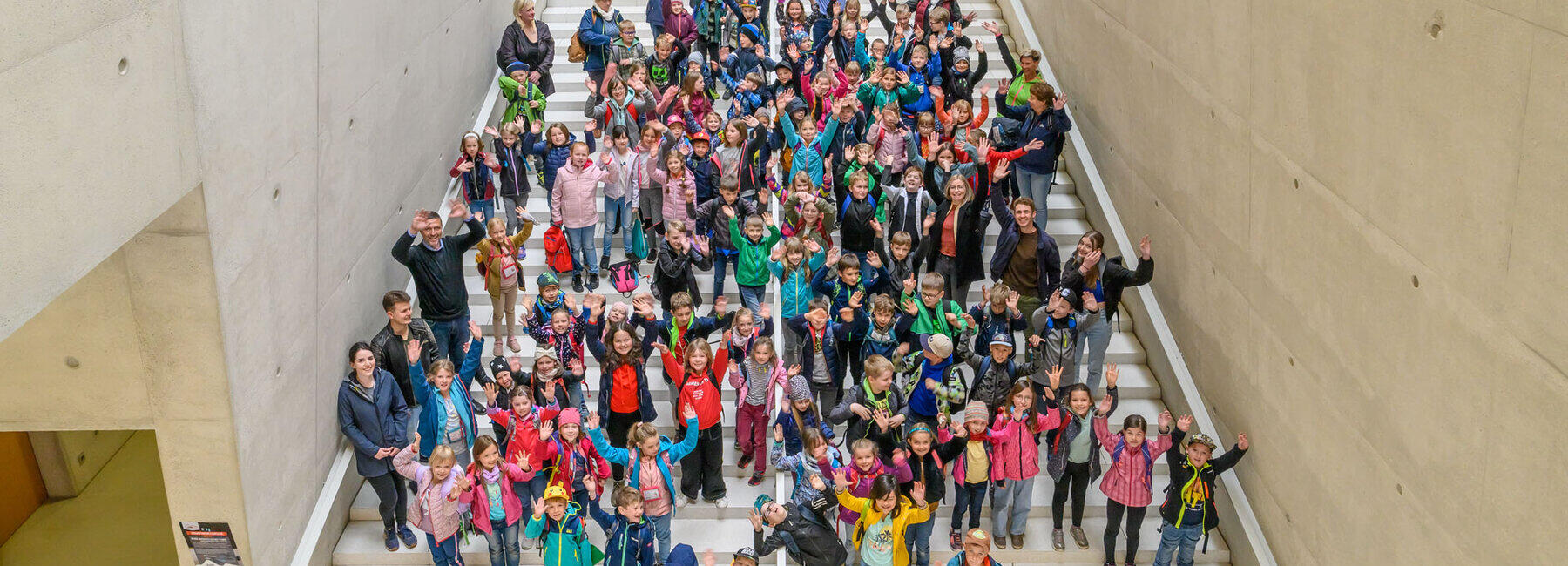 Blick von oben auf eine große Treppe. Viele Kinder und einige Erwachsene schauen mit hochgerissenen Armen jubelnd in die Kamera.