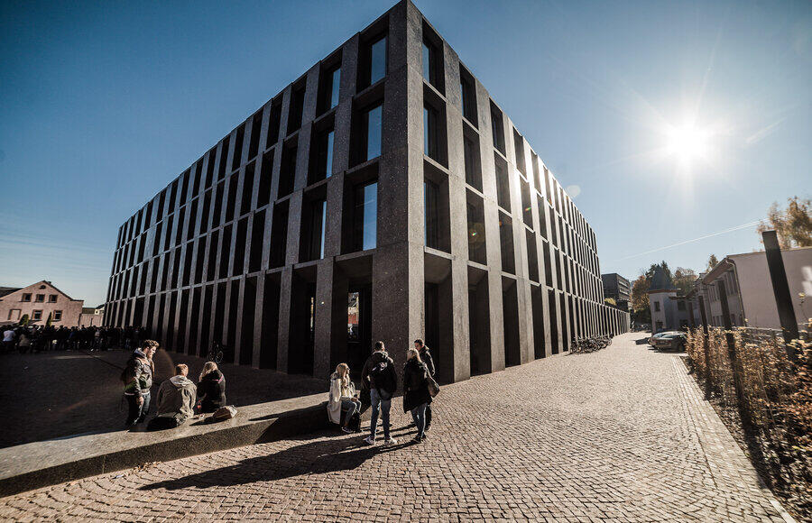 Modernes, mehrstöckiges Gebäude mit dunkler Fassade und vertikalen Fenstern an einem sonnigen Tag mit Menschen auf dem gepflasterten Vorplatz
