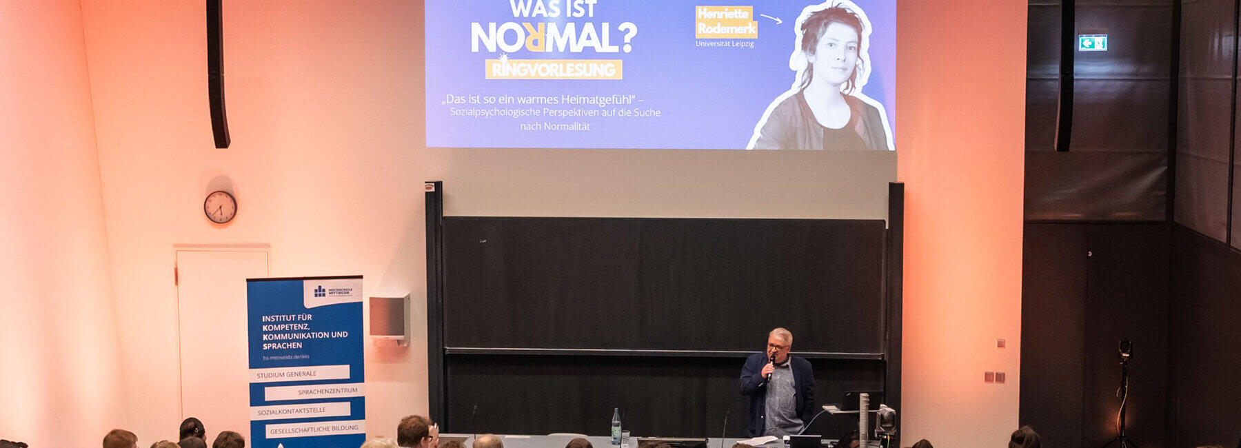 A view from the back, looking over the heads of the audience into the lecture theatre. Behind the lectern, a man is speaking to the audience, holding a microphone. On the left stands a blue roll-up banner displaying, amongst other things, the text ‘Institute for Competence, Communication and Languages (IKKS)’. The projection on the wall shows the welcome slide, featuring, amongst other things, the text ‘What is normal? Lecture series’ and the title of the first lecture: ‘That’s such a warm feeling of home’ – Social-psychological perspectives on the search for normality’. Next to it is a picture of the speaker, Henriette Rodemerk.