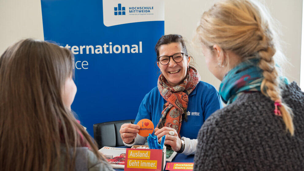Lachende weibliche Person vor "International-Office" Rollup hält ein "I love Erasmus+"-Kärtchen in den Händen und unterhält sich mit zwei Personen im Bildvordergrund