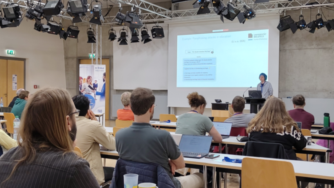 Person steht vor einer Präsentationswand, Zuhörende sitzen an Tischen