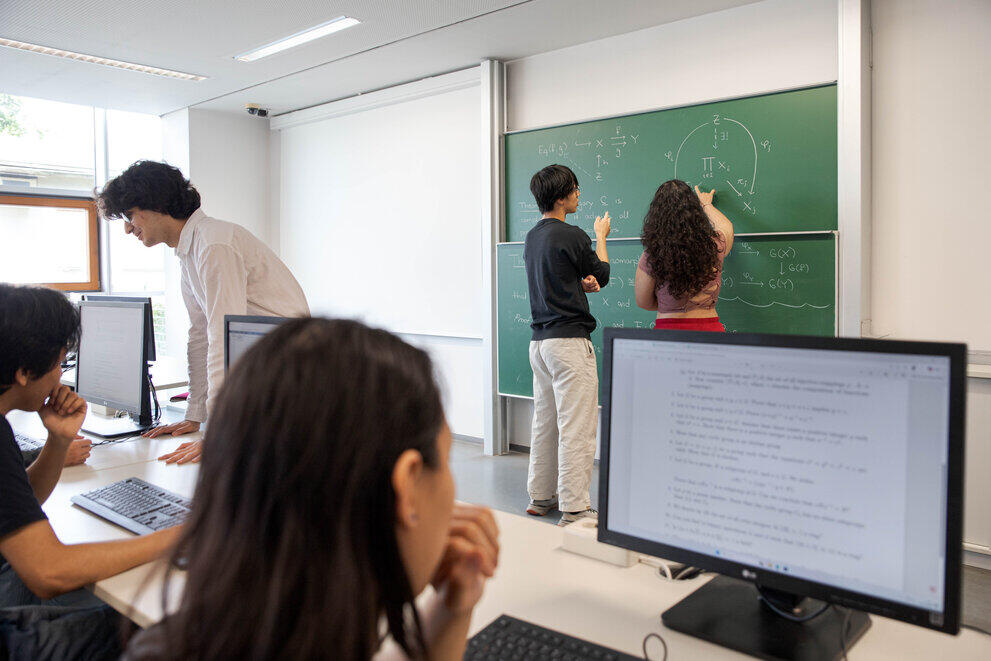 Zwei Studierende schreiben etwas an eine Tafel, während andere Studierende vor Computern sitzen.