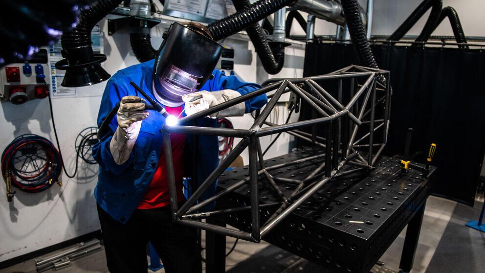 A person in overalls and a welding visor is welding a delicate frame made of metal rods.