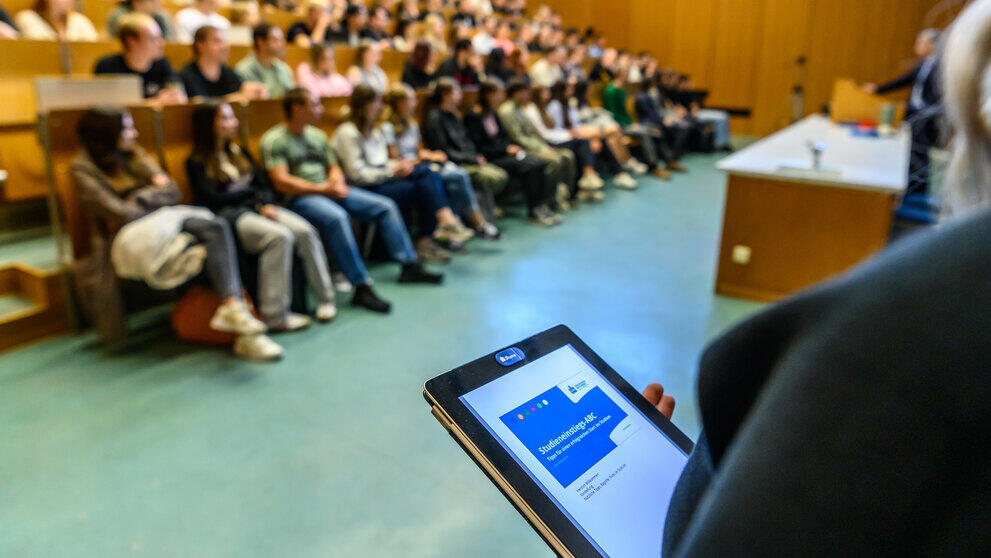 Blick über die Schultern einer weiblichen Person auf einen vollbesetzten Hörsaal mit ansteigenden Sitzreihen. Sie hät ein Tablet in der Hand, auf dessen Bildschirm Studieneingangs-ABC zu lesen ist.
