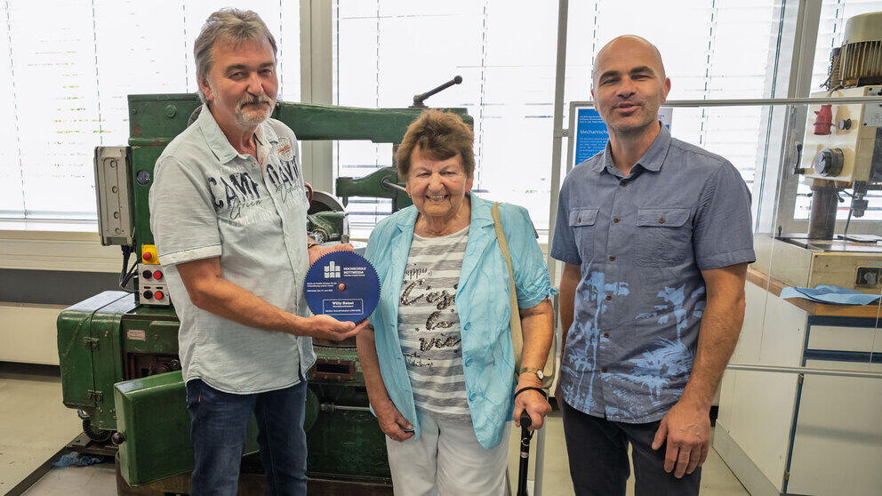 An elderly woman is standing in front of the machine, smiling, between two men. The man on the left is holding a large cogwheel in his hands, on which the Mittweida University of Applied Sciences logo can be seen against a blue background.