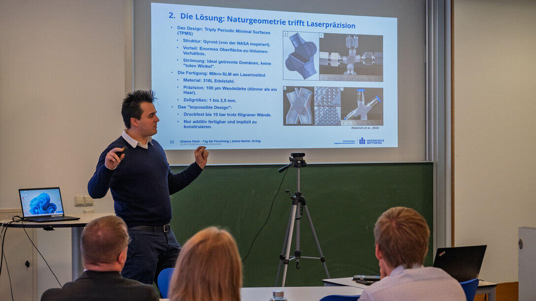 A man standing in front of a projected PowerPoint presentation. In the foreground, the backs of the audience’s heads.