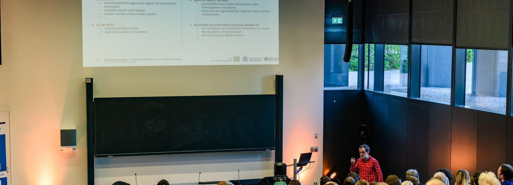 Blick von hinten in den Hörsaal über die Köpfe des Publikums auf den männlichen Dozenten. Schräg über ihm ist eine Folie projiziert mit der Überschrift: „Anwendungen von KI und maschinellem Lernen – III – ChatGPT & Co“ und „Was ist, was kann ChatGPT ¬ nicht?“