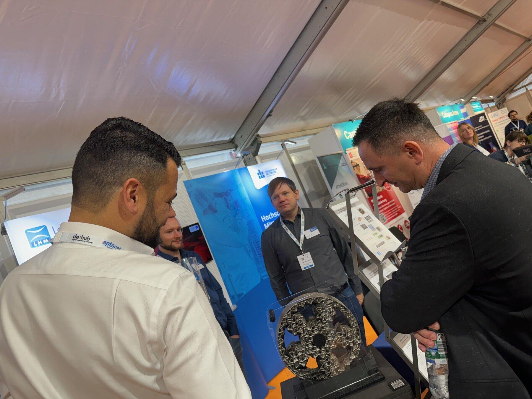 Drie männliche personen stehen auf einem Messestand und betrachten ein 3D-gedrucktes Planetengetriebe