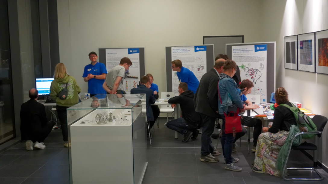 Besucher sitzen und stehen an verschiedenen Ausstellungstischen