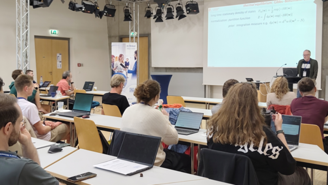 Person steht vor Präsentationswand, Zuhörende sitzen an Tischen