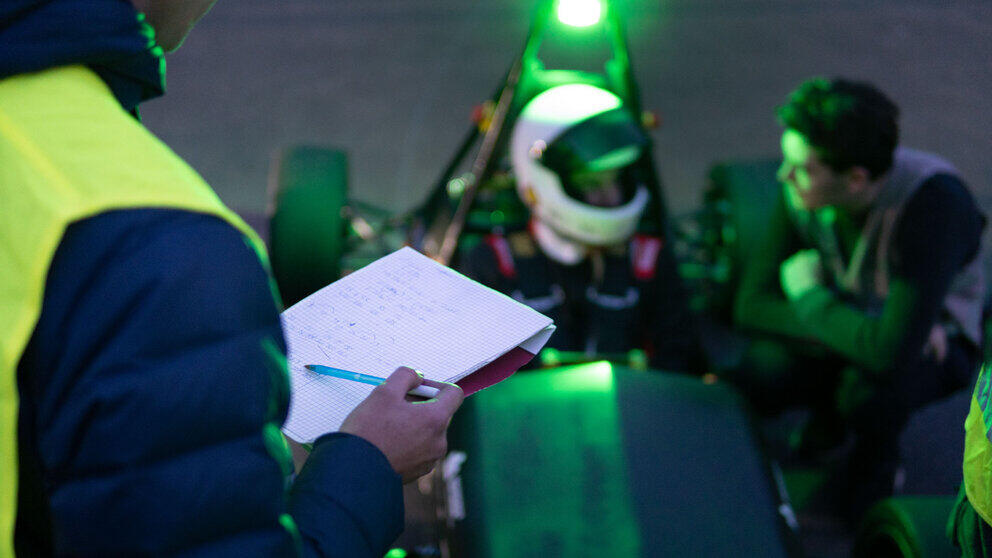 In the foreground of the picture, to the left, a person wearing a high-visibility vest and holding a notepad can be seen, shown from behind and partially in profile. In the background, the driver in a racing car wearing a helmet is visible out of focus, and next to him a man is kneeling; the two are seen talking to one another.