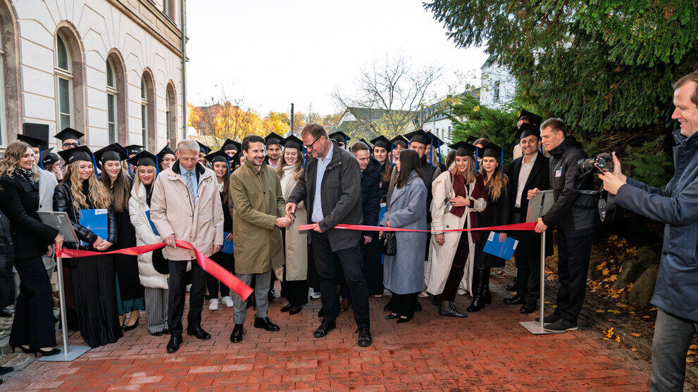 Three men are cutting a red ribbon on a paved path. Behind them stand numerous graduates.