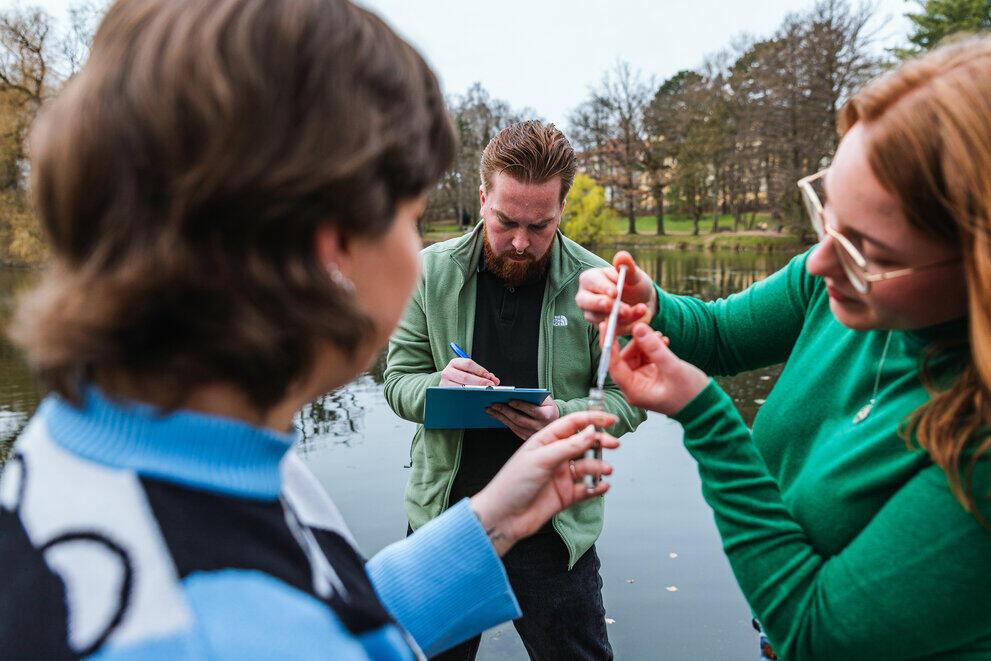Three students are standing by a pond, filling a test tube with a water sample.