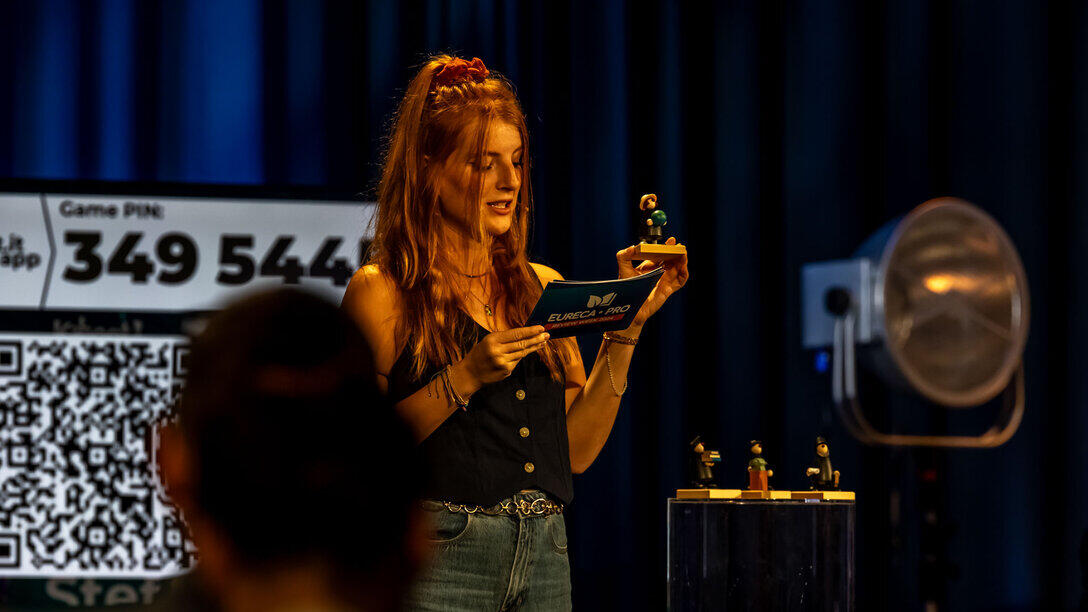A presenter in a television studio is hosting a quiz. She is holding a small wooden figurine, which is the prize awaiting the winner.