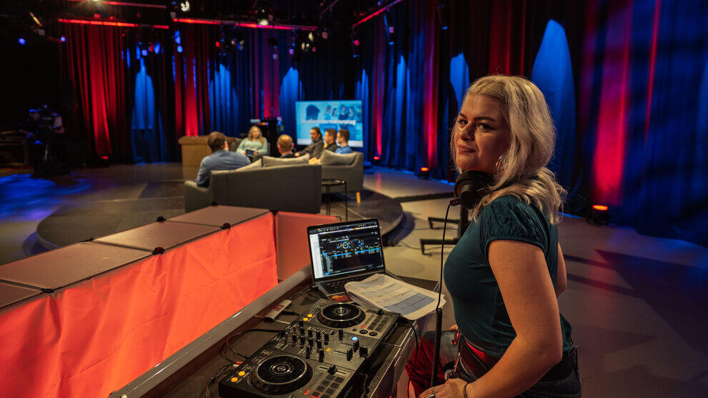 A young woman at the DJ desk in the TV studio. In the background, the people on the podium.