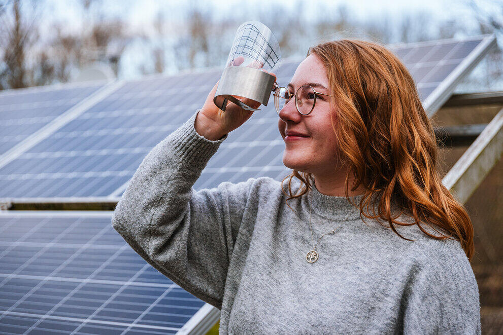 Eine Studentin steht vor Photovoltaik-Paneelen und schaut durch ein Messgerät in den Himmel.