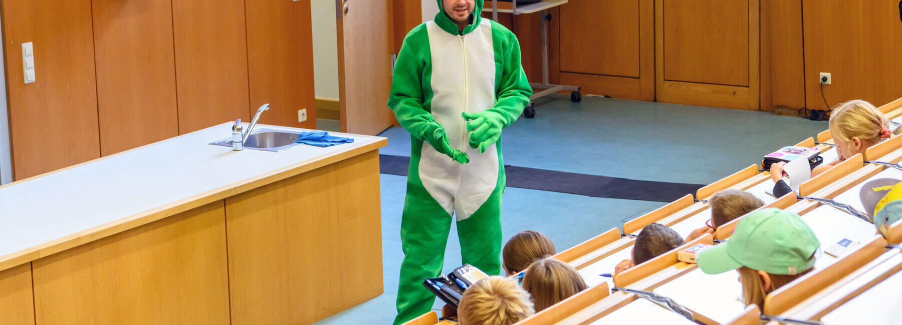 Ein freundlicher Mann im grünen Ganzkörper-Froschkostüm steht vor ienem Pult im Hörsaal. Im Bildvordergrund sitzen Kinder in den ansteigenden Bankreihen.