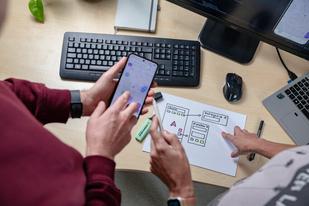 Two students are standing by a desk, pointing at a smartphone.