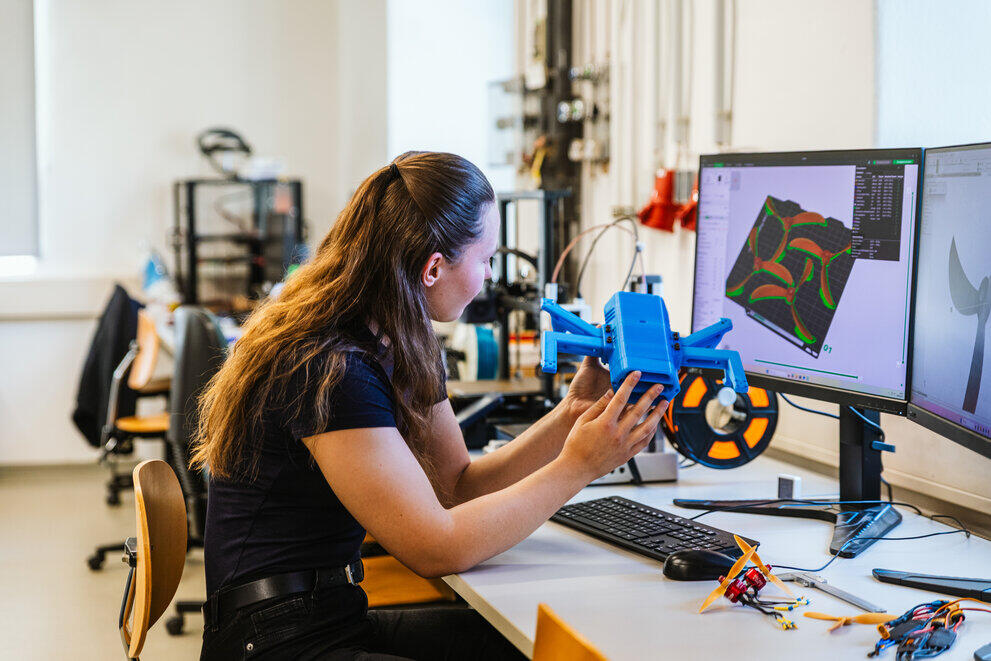 Eine Studentin hält eine 3D-gedruckte Drohne und vergleicht sie am PC mit der CAD-Konstruktion.