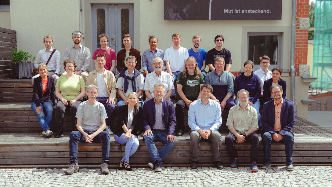 24 Personen sitzen für eien Gruppenfoto auf einem ansteigenden Podest aus Holz vor einer Hauswand