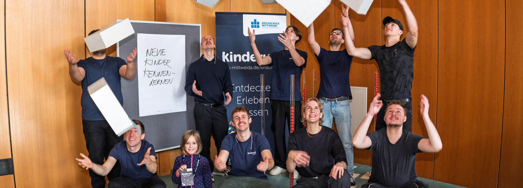 Eine Gruppe von zehn Personen im Hörsaal, die Schubladen aus Karton in die Luft werfen und wieder auffangen. Im Hintergrund sieht man ein dunkelblaues Kinderuni-Banner und eine Stellwand mit der Aufschrift „Neue Kinder kennenlernen“.