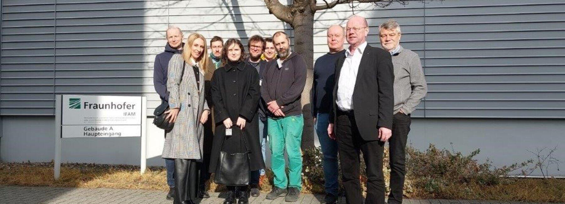 A group of 10 people are standing in front of a house wall and next to a Fraunhofer IFAM sign