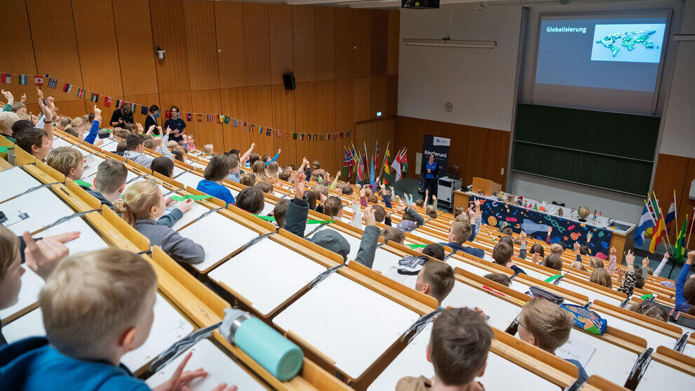 Großer Hörsaal mit vielen Kindern, die auf eine Präsentation zum Thema 'Globalisierung' auf einer Leinwand schauen. Viele Nationalflaggen als Wimpel an den Wänden und auf Fahnenständern am Fuß des Hörsaals.