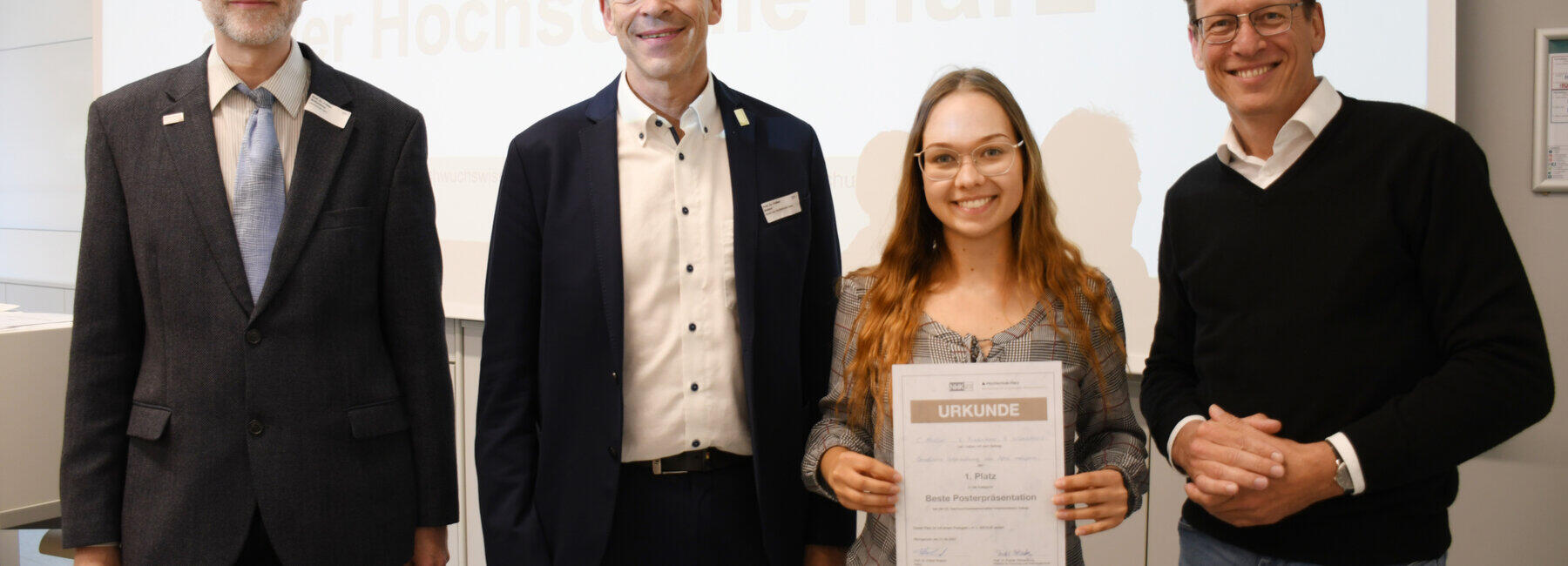 Drei männliche Personen und eine weibliche Person stehen vor einem beleuchteten Hintergrund, die weibliche Person hält eine Urkunde in den Händen