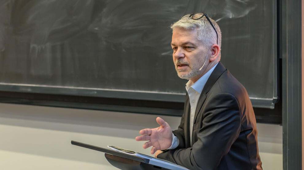 Eine männliche Person mit entschiedener Mine vor einer schlecht geputzten Tafel, die Brille nach oben ins Haar geschoben, den linken Ellbogen auf ein Rednerpult abgestützt und die rechte Hand geöffnet, spricht zu einem im Bild nicht sichtbaren Publikum.