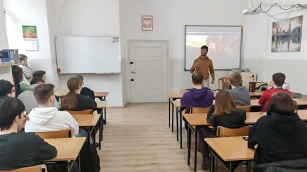 Ein junger Man seht in einem Seminarraum vor einer Leinwand, an die ein Bild projiziert ist, das nicht erkennbar ist. Mehrere Personen an Tischen hören zu.