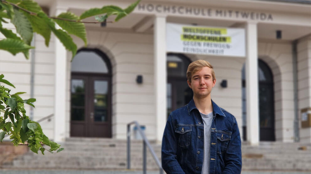 Ein junger Mann steht vor einem Haus mit einem Transparent "weltoffene Hochschule".