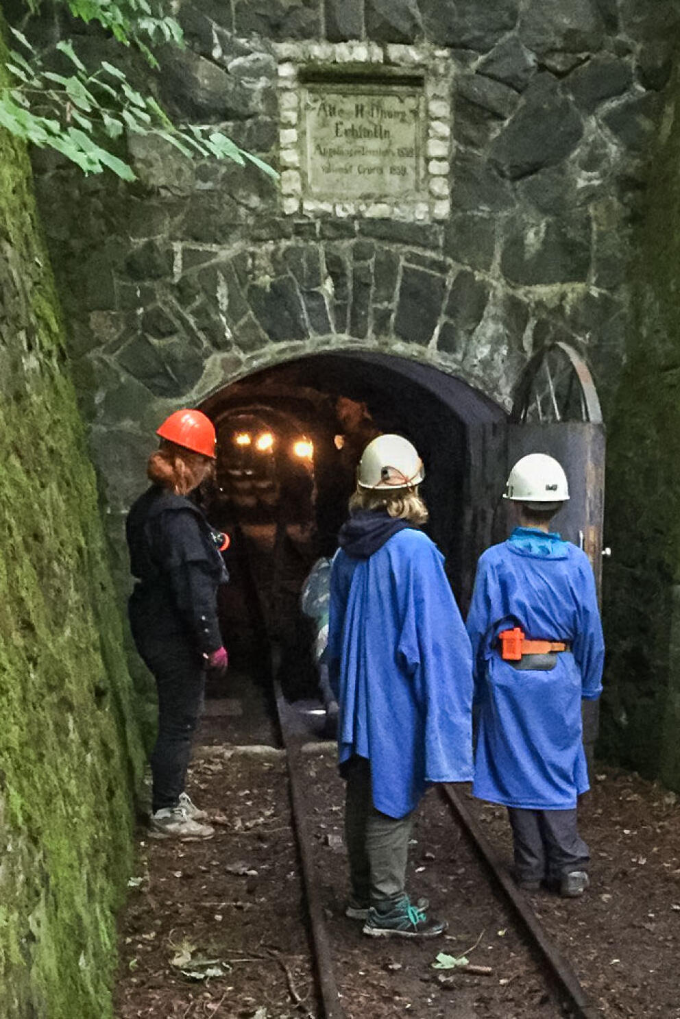 Drei Kinder stehen mit Schutzhelm und Regenjacke vor dem Eingang zu einem Stollen. Bahngleise führen in den Stollen. Die Tür ist geöffnet, gibt den Blick ins Innere frei.