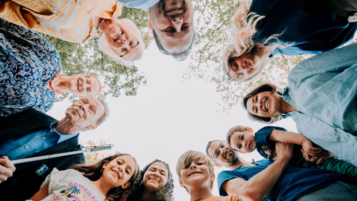 Eleven people of different ages, genders and skin colours are standing in a circle, looking down at the camera. Above them, in the centre, there is an unobstructed view of the sky.