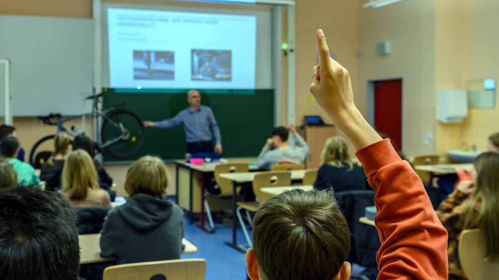 Blick von hinten in einen Klassenraum über die Köpfe der Schüler. Im Hintergrund steht ein männliche Person vor einer Tafel. Im Bildvordergrund ist der zum Melden gehobene Arm eines Schüler zu sehen.