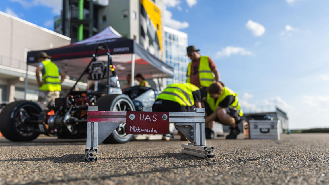 Ein Schild mit dem handgeschriebenen Text „UAS Mittweida“ steht auf dem Boden vor einem Rennwagen, an dem Personen hantieren. Im Hintergrund ist unscharf ein hohes Gebäude vor blauem Himmel zu sehen.