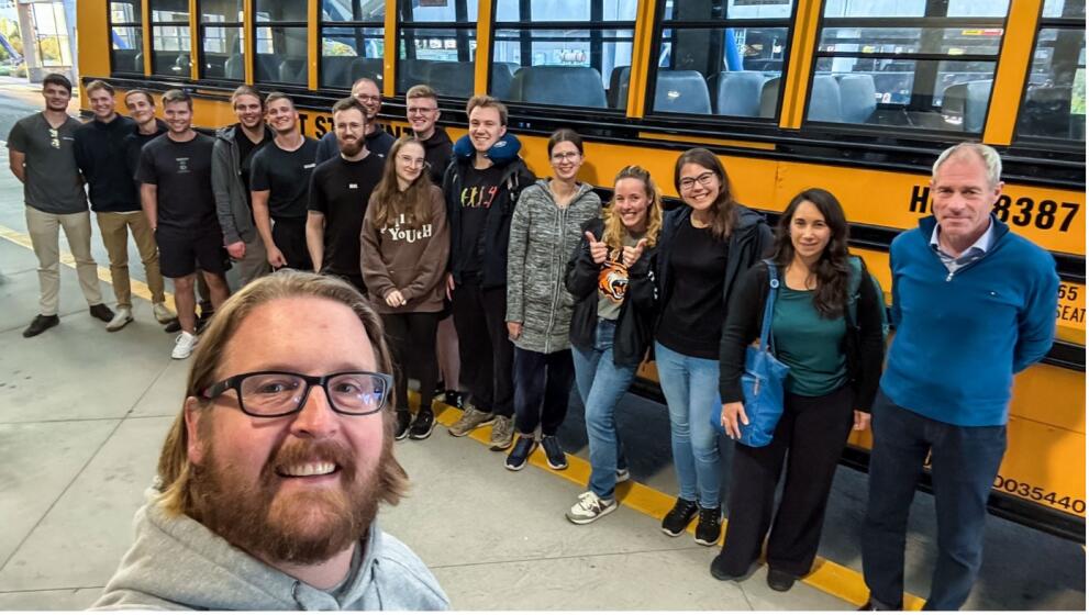 The selfie shows a group of mostly young people in front of a typical yellow American school bus. The photographer’s head can be seen in the foreground of the picture.