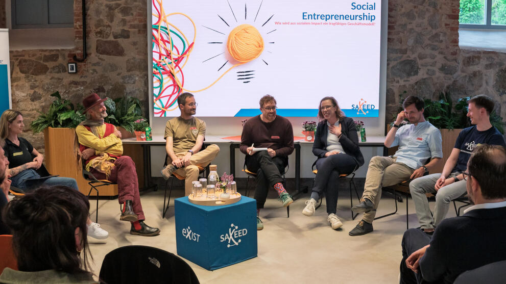 Sieben Personen auf Stühlen vor einem großen Screen, auf dem „Social Entrepreneurship. Wir wird aus sozialem Impact ein tragfähiges Geschäftsmodell“ zu lesen ist. Im Bildvordergrund sind Köpfe von Zuschauenden von hinten zu sehen.