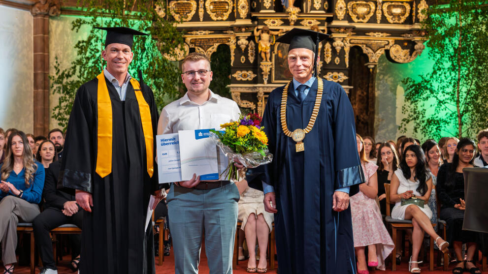 Three men standing in the chancel of a church. The younger man in the middle is holding a bouquet of flowers and a certificate. The men on the left and right are wearing academic gowns and caps; the man on the right is also wearing a gold chain of office.