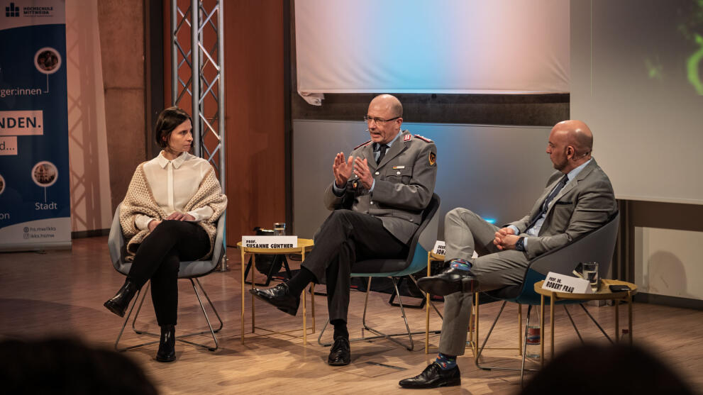 One woman and two men sitting in armchairs arranged in a semicircle. The person in the middle is wearing a Bundeswehr uniform.