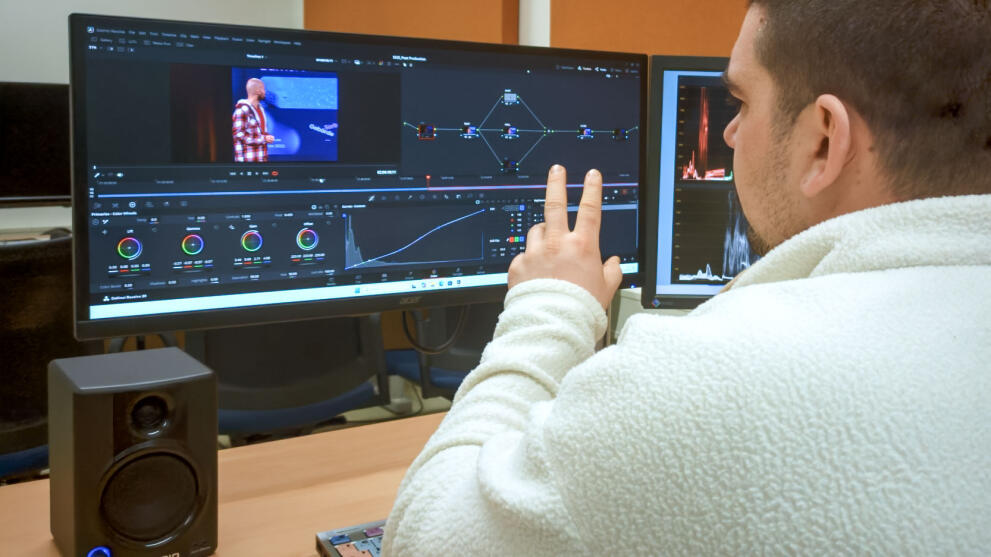 A close-up shot of a person sitting at a desk from behind, in front of a large monitor.