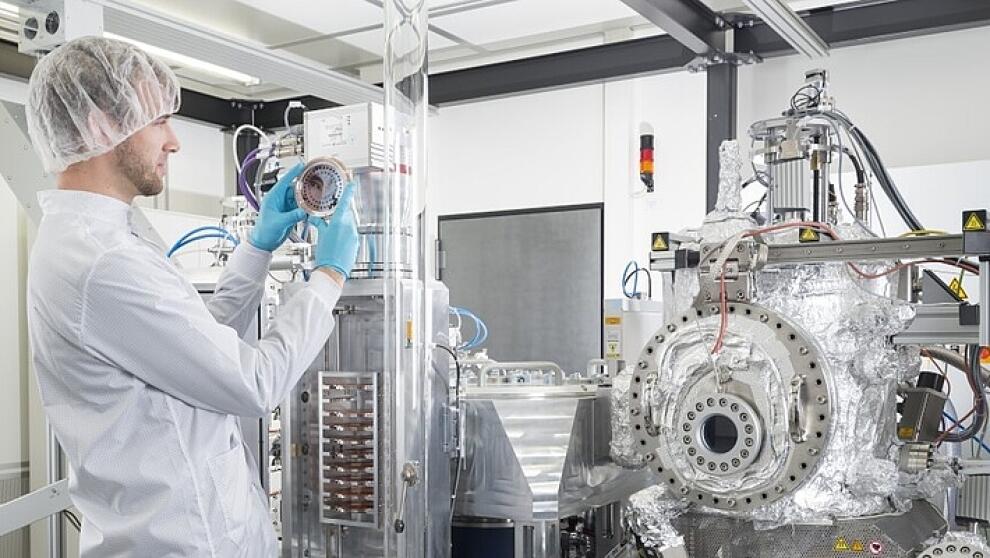 Männliche Person in Reinraumschutzkleidung steht vor Anlage und hält einen Wafer in der Hand