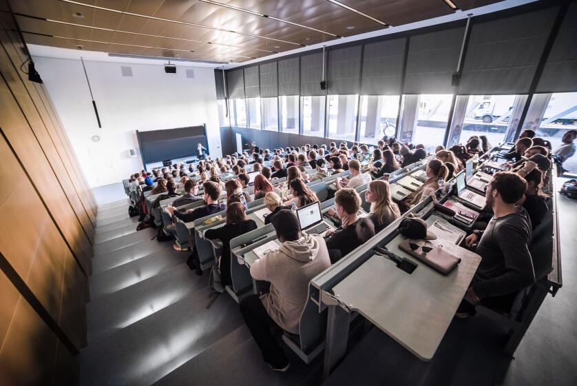Hörsaal mit vielen Studierenden, die auf Reihenplätzen sitzen und einem Dozenten zuhören, der vor einer Tafel steht