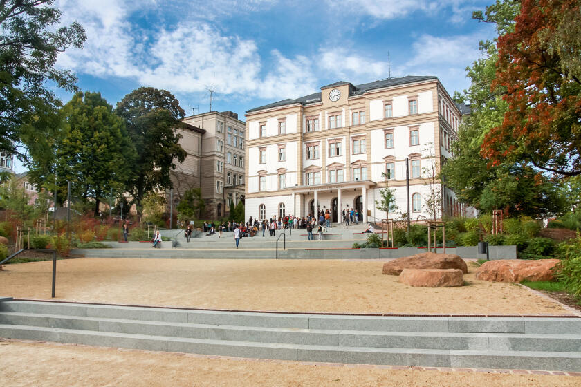 Großes historisches Gebäude mit mehreren Stockwerken, davor ein offener Platz mit Bäumen und Bänken
