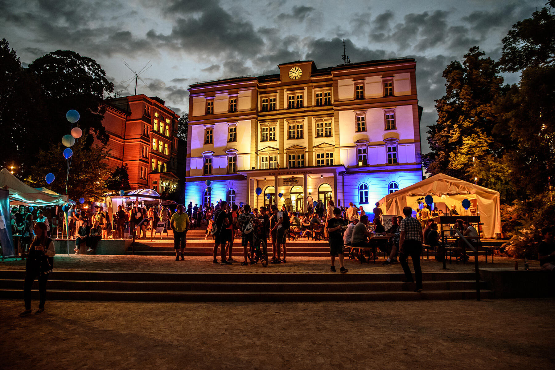 Das Hauptgebäude der Hochschule Mittweida leuchtet bunt zur Nacht der Wissenschaften. Davor sind Stände platziert.