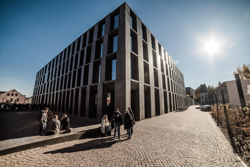 Modernes, mehrstöckiges Gebäude mit dunkler Fassade und vertikalen Fenstern an einem sonnigen Tag mit Menschen auf dem gepflasterten Vorplatz