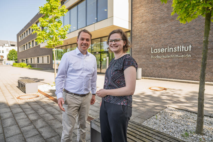Eine männliche udn eine weibliche Person stehen nebeneinander und gestikulieren freundlich. Im Hinterggrund ist iein verklinkertes Gebäude mit dem Schriftzug "Laserinstitut Hochschule Mittweida" zu sehen.
