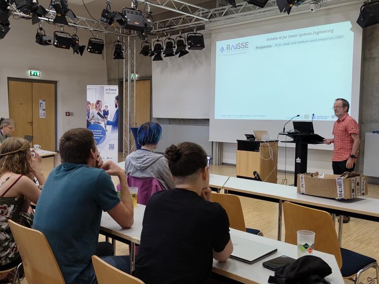 Person steht vor Präsentationswand, Zuhörende sitzen an Tischen